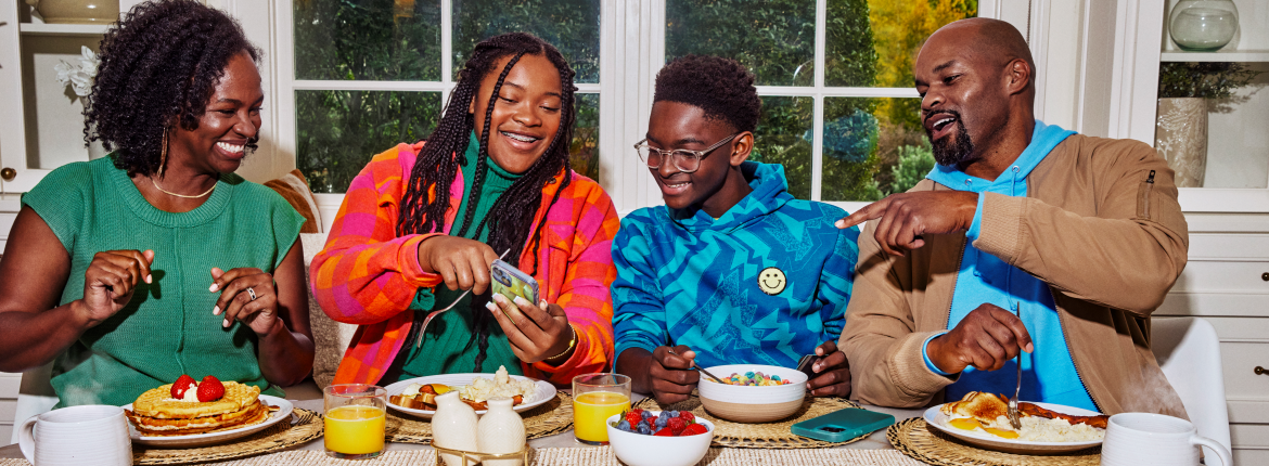 a family seated at a table sharing a meal; the daughter is sharing her smartphone screen with the others